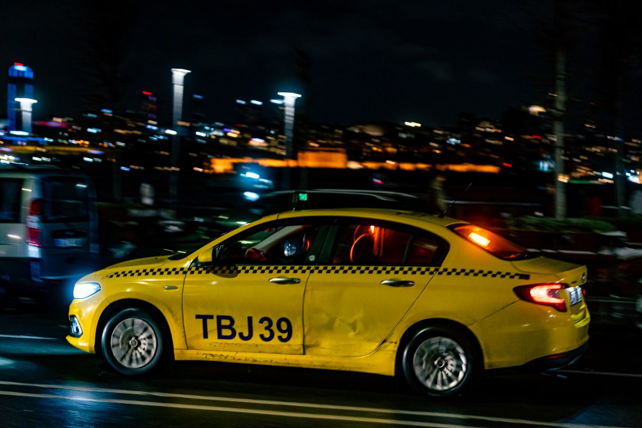 hero-img-01 Vivid night street scene with a yellow taxi driving through city streets, showcasing urban nightlife.