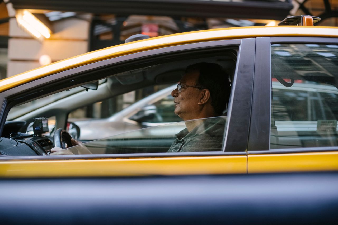 services-01 Side view of ethnic male driver sitting behind steering wheel driving yellow cab with lowered window on city in street with blurred background